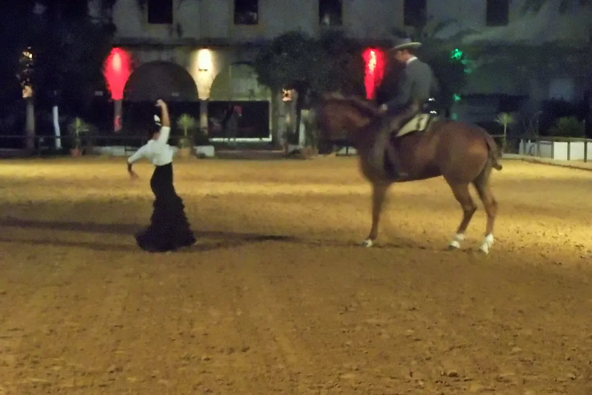 Eine Flamenco-Tänzerin in einem schwarzen Kleid hebt einen Arm neben einem Reiter auf einem kastanienbraunen Pferd in einer flutlichtbeleuchteten Sandarena bei Nacht, dahinter ein beleuchtetes Steingebäude