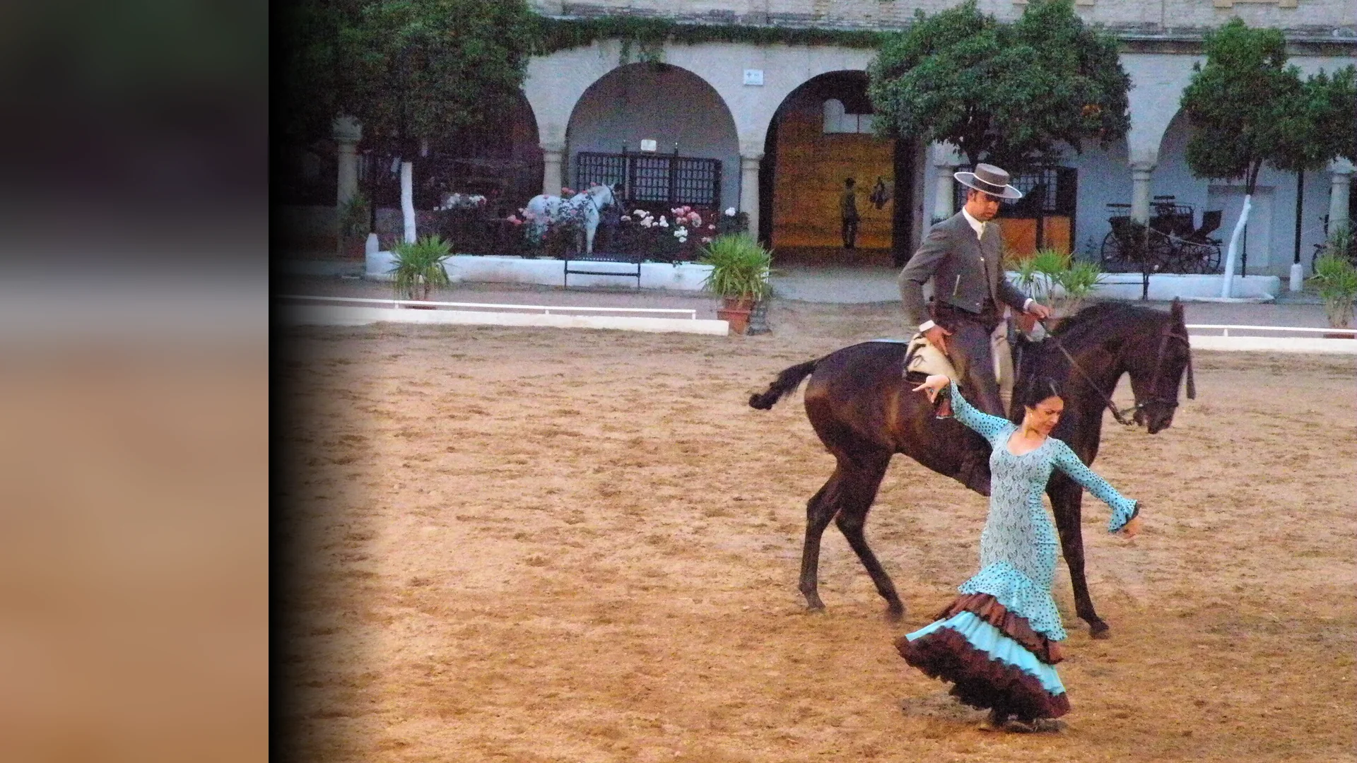 Tanečnice flamenca v tyrkysových volánových šatech se otáčí vedle jezdce na tmavém hnědákovi v pískové aréně, za nimi bílé arkády a pomerančovníky.